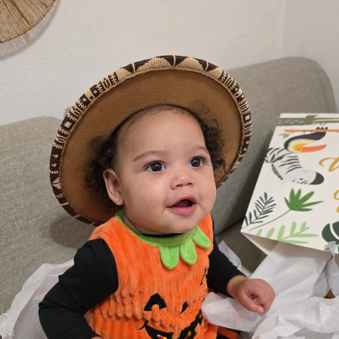 A baby wearing a pumpkin costume with an intricately beaded hat by Oklahoma Native bead artist, sitting on a gray couch next to a gift box.