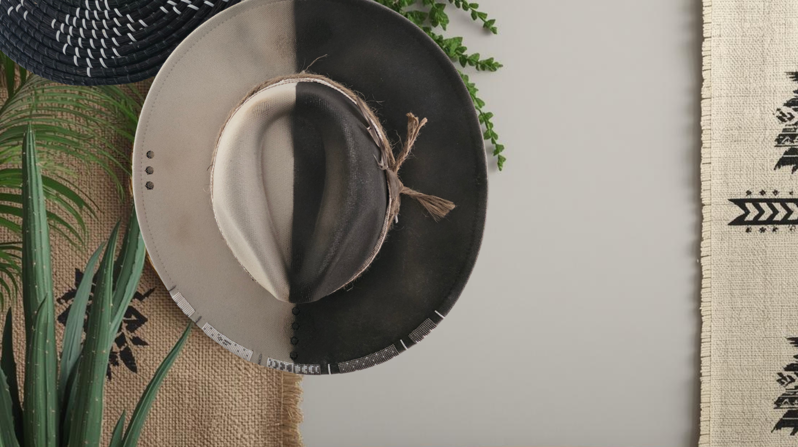A color block half matte black and half warm beige wide brim  mens fedora hat, styled on a neutral background with decorative plants, a woven mat, and a patterned textile.
