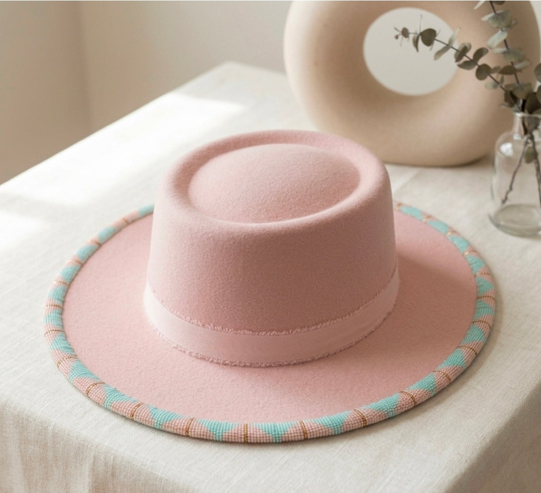A light pink felt wide-brim hat with a matching pink band and a striped brim edge in pink, beige, and light blue, displayed on a white tablecloth.