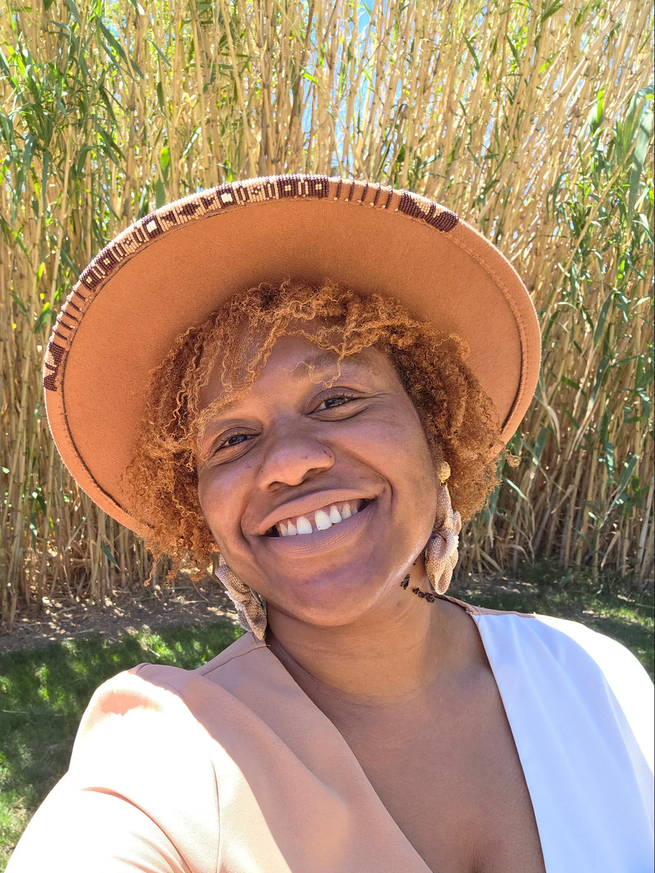 A smiling woman outdoors wearing a wide-brimmed tan hat with southwestern plains tribe inspired beaded hat brim, with tall bamboo in the background.